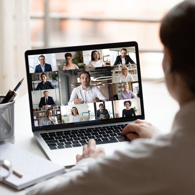 View over businesslady shoulder seated at workplace desk look at computer screen where collage of many diverse people involved at video conference negotiations activity, modern app tech usage concept